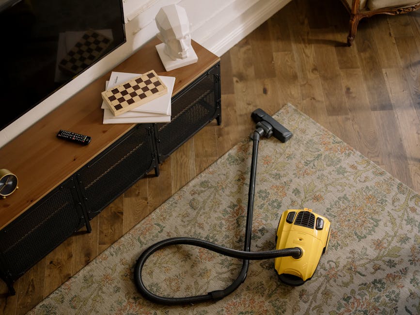 A yellow vacuum cleaner with a black hose and metal wand attachment is positioned on a floral-patterned area rug in a living room, near a wooden sideboard with metal legs. The sideboard holds a white 