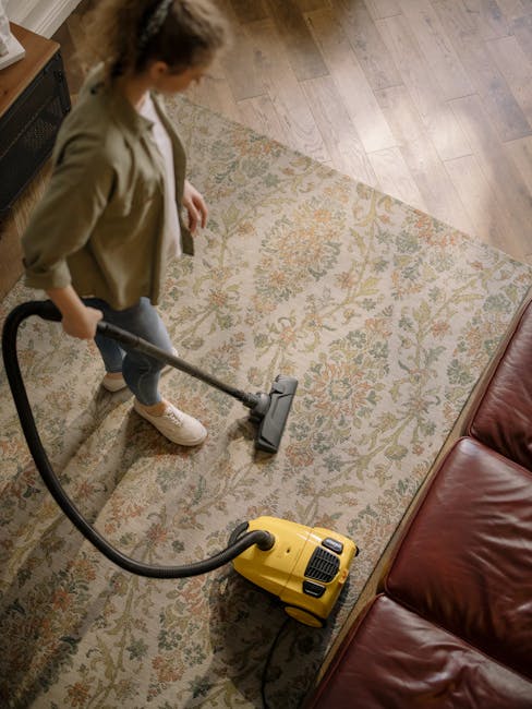 A yellow vacuum cleaner with a black hose and metal wand attachment is positioned on a floral-patterned area rug in a living room, near a wooden sideboard with metal legs. The sideboard holds a white 